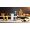 Na zdjęciu znajduje się sokowirówka CECOTEC Juice Fresh 400 Titan na blacie kuchennym, obok której stoją dwa szklanki z zielonym sokiem oraz dzbanek z sokiem. W tle widoczne są różne warzywa, deska do krojenia i drewniana deska dekoracyjna.