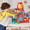 Na zdjęciu znajduje się dziecko bawiące się zestawem figurek przedstawiającym siedzibę Iron Mana z serii Disney Jr. Marvel. Zestaw jest kolorowy, z dużą maską Iron Mana na górze oraz różnymi figurkami i elementami do zabawy.