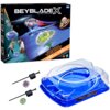 Na zdjęciu znajduje się opakowanie zestawu "Beyblade X" z napisem "Drop Attack" oraz ilustracjami wirujących zabawek i dwóch osób. Obok opakowania widoczna jest niebieska arena do gry oraz dwa wirujące bączki z wyrzutniami.