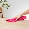 Różowa ściereczka z napisem "VELKEA HOME SMART FIBRE" jest używana do wycierania drewnianej powierzchni. W tle widoczna jest roślina w doniczce na drewnianym stojaku.