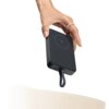 Na zdjęciu widoczny jest szary powerbank z okrągłym symbolem błyskawicy na powierzchni, trzymany w dłoni. Powerbank ma porty i wskaźniki LED na krawędzi oraz podłączony jest do niego kabel.