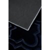 Fragment granatowego dywanu z widocznym geometrycznym wzorem oraz rogiem odwróconym, ukazującym czarną, antypoślizgową powierzchnię z białymi kropkami. Krawędzie dywanu są obszyte jasną nicią.
