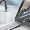 Na zdjęciu widać czarny kabel USB-C podłączony do laptopa oraz białej ładowarki z niebieskim podświetleniem. Na ekranie laptopa widoczne są ikony aplikacji oraz tapeta z krajobrazem górskim.