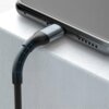 Czarny kabel USB-C jest podłączony do urządzenia leżącego na jasnej powierzchni, z widocznymi detalami konstrukcji kabla, takimi jak niebieskie linie na zgięciu. Na wtyczce kabla znajduje się logo producenta.