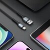 Na zdjęciu znajduje się czarny kabel USB-Lightning z metalowymi końcówkami, ułożony na ciemnym tle, otoczony przez fragmenty trzech smartfonów. Kabel ma plecioną osłonę, a na jednej z końcówek widnieje logo "3MK".