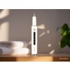 Na zdjęciu znajduje się skaler do zębów ALPHA DentiPro AWS-03 ustawiony pionowo na drewnianej powierzchni, obok dwóch złożonych ręczników. W tle widoczne są rozmyte liście rośliny, a na produkcie oraz w prawym dolnym rogu zdjęcia znajduje się napis "DentiPro".