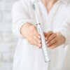Osoba trzyma białą szczoteczkę soniczną z widocznym napisem "DentiPro" na uchwycie. Na szczoteczce znajdują się przyciski oraz oznaczenia trybów: Clean, White, Polish, Massage.