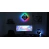 Na zdjęciu znajduje się biurko z monitorem wyświetlającym kolorową grę, klawiaturą i myszką, a na ścianie wisi neon LED z napisem "GAMER ZONE" oraz grafiką kontrolera, emitujący wielobarwne światło. Obok monitora znajduje się stojak na słuchawki z delikatnym podświetleniem, a na półce widoczne są pudełka z grami.
