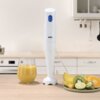 Na zdjęciu znajduje się biały blender ręczny z niebieskim przyciskiem, obok którego stoi szklanka z żółtym napojem oraz talerz z pokrojonymi owocami, takimi jak kiwi, pomarańcza, limonka i mango. Na blenderze widoczne jest logo "BRAUN".