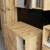 Zestaw mebli łazienkowych wiszących wykonany z drewna w kolorze dąb artisan, z widocznymi otworami na blacie oraz czarnym, marmurowym panelem na ścianie. Meble posiadają szuflady i półki, na których znajdują się różne przedmioty.