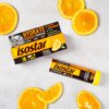Na zdjęciu widoczny jest produkt "Isostar Hydrate & Perform Powertabs" o smaku pomarańczowym, w postaci opakowania kartonowego oraz tuby, otoczony plasterkami pomarańczy. Na opakowaniu znajdują się napisy: "Hydrate & Perform Powertabs", "Orange Flavour", "500 ml", oraz logo "Isostar".