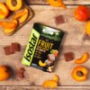 Opakowanie żelków "Isostar Fruit Boost Energy" o smaku morelowym leży na drewnianym blacie, otoczone kawałkami świeżych moreli i pojedynczymi żelkami. Na opakowaniu widnieje napis: "FRUIT BOOST ENERGY with caffeine", a także informacje o zawartości witamin i ilości żelków (10 sztuk).