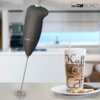 Na zdjęciu znajduje się czarny spieniacz do mleka z logo "CLATRONIC" oraz szklanka z napisem "Kaffee Coffee Caffe" wypełniona napojem, stojąca na talerzyku z dwoma ciastkami i łyżeczką. W tle widoczna jest rozmyta kuchnia.