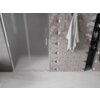 Na zdjęciu widoczne są drzwi prysznicowe z matowego szkła z chromowanym uchwytem, obok których znajduje się ściana wyłożona płytkami o geometrycznym wzorze, na której wisi biały ręcznik. W tle widać wnękę z półkami, na których stoją różne butelki.