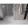 Na zdjęciu widoczne są drzwi prysznicowe z matowego szkła z białą ramą i pionowym uchwytem, obok znajduje się ściana wyłożona płytkami w geometryczny wzór, na której wisi ręcznik. W tle widać wnękę z półkami, na których stoją różne butelki.
