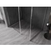 Przesłane zdjęcie przedstawia fragment łazienki z prysznicem typu walk-in, wyposażonym w przezroczystą ściankę z chromowaną ramką. Podłoga i ściany są wyłożone ciemnymi płytkami o wzorze jodełki, a obok prysznica stoi mały stolik z butelką.