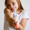 Na zdjęciu widoczna jest biała szczoteczka soniczna w kształcie kota, trzymana w dłoni, z podświetlaną końcówką. W tle znajduje się osoba w białej koszulce, która się uśmiecha.