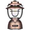 Latarka biwakowa w stylu retro z metalicznym wykończeniem, posiada przezroczysty klosz z widocznymi żarówkami LED oraz pokrętło do regulacji światła. Na podstawie znajduje się logo "OLIGHT".