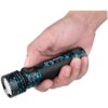Osoba trzyma w dłoni latarkę z czarno-niebieskim wzorem, na której widoczne są napisy "OLIGHT" i "Seeker 4 Pro". Latarka ma okrągły przycisk z symbolem słońca.