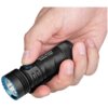 Na zdjęciu widoczna jest czarna latarka trzymana w dłoni, z niebieskim przyciskiem i napisem "OLIGHT Seeker 4 Mini" na obudowie. Latarka ma żebrowaną powierzchnię i niebieską krawędź wokół reflektora.