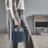 Osoba trzyma w ręce niebieski grzejnik elektryczny z dwoma pokrętłami i widocznym logo "ROWENTA". W tle znajduje się szafka z drewnianym uchwytem.