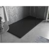 Czarny, prostokątny brodzik prysznicowy o wymiarach 160 x 70 cm znajduje się na tle ściany wyłożonej szarymi, mozaikowymi płytkami. Na pierwszym planie widoczny jest fragment okrągłego stolika z butelkami, a podłoga jest wykonana z szarego materiału.