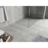 Szary brodzik prostokątny zintegrowany z podłogą w łazience, otoczony dużymi płytkami ceramicznymi w podobnym odcieniu. Na pierwszym planie widoczny jest fragment drewnianej półki z kosmetykami oraz ręcznik wiszący na ścianie.
