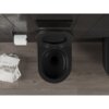 Czarna matowa miska WC typu Rimless Tornado jest zamontowana na ciemnej ścianie, obok znajduje się uchwyt z rolką papieru toaletowego oraz stos magazynów. Na podłodze widoczne są drewniane panele, a na ścianie przycisk spłukujący.