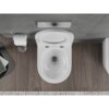 Biała, połyskująca miska WC typu Rimless Tornado z deską wolnoopadającą slim, wykonana z duroplastu, zamontowana na tle szarej ściany i drewnianej podłogi. Obok znajduje się uchwyt z rolką papieru toaletowego, a na podłodze leży dywanik z jasnymi frędzlami.