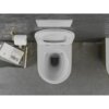 Biała miska WC z deską wolnoopadającą, widziana z góry, zamontowana na szarej ścianie. Obok znajduje się uchwyt na papier toaletowy oraz stos książek, z widocznym tytułem "John Ruskin VON Venedig".