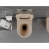 Na zdjęciu widoczna jest miska WC w kolorze cappuccino mat z deską wolnoopadającą, umieszczona na tle szarej, betonowej ściany. Obok znajduje się uchwyt z rolką papieru toaletowego oraz stos książek, z których jedna ma widoczny tytuł "John Ruskin Von Venedig".