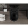 Czarna matowa miska WC z deską wolnoopadającą znajduje się na tle ciemnych płytek ściennych i drewnianej podłogi. Obok widać uchwyt z rolką papieru toaletowego oraz stos magazynów, z widocznym napisem "sofa" na okładce.