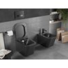 Na zdjęciu znajduje się czarna matowa miska WC typu rimless oraz bidet, zamontowane na ścianie wyłożonej szarymi płytkami. Obok miski WC wisi uchwyt z papierem toaletowym, a na półce znajdują się książki i rośliny.