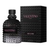 Czarna butelka perfum o fakturze przypominającej piramidy, z różowym napisem "VALENTINO" na etykiecie, obok czarnego pudełka z różowym napisem "VALENTINO UOMO" oraz "BORN IN ROMA". Na dole pudełka widnieje napis "EAU DE TOILETTE VAPORISATEUR SPRAY 100 ml 3.4 FL.OZ.".