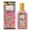 Różowa butelka perfum z złotym korkiem obok ozdobnego pudełka, oba z motywem kwiatowym. Na obu widnieje napis: "GUCCI FLORA GORGEOUS GARDENIA", a na pudełku dodatkowo: "Eau de Parfum Vaporisateur Natural Spray 50 ml 1.6 FL OZ".