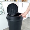 Czarny plastikowy kosz na śmieci o pojemności 47 litrów z otwartą pokrywą o strukturze plastra miodu, obok którego widoczna jest ręka naciskająca przycisk. W tle znajduje się biała szafka kuchenna z uchwytami oraz wiszący ręcznik i ściereczka z roślinnym wzorem.