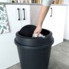 Czarny plastikowy kosz na śmieci o pojemności 47 litrów z uchylną pokrywą, do którego wkładana jest biała plastikowa torba. W tle widoczna jest biała szafka kuchenna z czarnymi uchwytami oraz blatem, na którym stoją pojemniki z produktami spożywczymi.