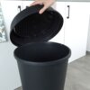 Czarny plastikowy kosz na śmieci o pojemności 47 litrów z otwartą pokrywą, trzymaną przez rękę, znajduje się na tle białych szafek kuchennych. Podłoga jest wykonana z szarego materiału, a obok widoczna jest zasłona z roślinnym wzorem.