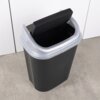 Czarny plastikowy kosz na śmieci o pojemności 25 litrów z szarym uchylnym wiekiem, ustawiony na jasnej podłodze przy białej ścianie. Kosz ma prostokątny kształt i gładką powierzchnię.