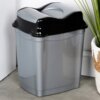 Na zdjęciu znajduje się szary plastikowy kosz na śmieci o pojemności 16 litrów, z czarną pokrywą. Kosz stoi na podłodze obok białej ściany i rośliny w plecionym koszu.