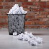 Czarny, metalowy kosz na śmieci wypełniony zgniecionymi kartkami papieru stoi na tle ceglanej ściany. Na podłodze leży kilka zgniecionych kartek papieru.