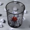 Czarny, metalowy kosz na śmieci o wymiarach 26x28 cm jest wypełniony zgniecionymi kartkami papieru, w tym jedną czerwoną. Wokół kosza leżą dodatkowe zgniecione kartki papieru na szarej podłodze.