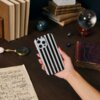 Na zdjęciu widać rękę trzymającą telefon w etui z czarno-białymi pionowymi paskami, na drewnianym stole leżą książki, lupa, kartka z nutami oraz szklana kula. W tle znajdują się stosy książek i dekoracyjne kule.