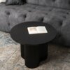 Okrągły, czarny stolik kawowy o ryflowanej podstawie stoi na wzorzystym dywanie, a na jego blacie leży otwarta książka. W tle widoczna jest szara, pikowana sofa.