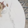 Okrągły stolik kawowy z białym marmurowym blatem, na którym stoi biały wazon z rośliną. W tle widoczny jest wzorzysty dywan w odcieniach szarości i beżu.