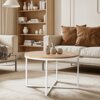Okrągły stolik kawowy o skandynawskim designie z białym metalowym stelażem i blatem w kolorze dębu, na którym stoją trzy ceramiczne wazony. W tle znajduje się jasna sofa z brązowymi poduszkami oraz regał z książkami i dekoracyjnymi wazonami.