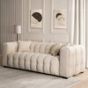 Kremowa, trzyosobowa sofa o pikowanej powierzchni stoi na jasnym dywanie w eleganckim salonie z jasnymi zasłonami i dwoma abstrakcyjnymi obrazami na ścianie. Obok sofy znajduje się lampa stojąca z białym abażurem.