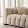 Kremowa, trzyosobowa sofa o pikowanej powierzchni z dwoma poduszkami, ustawiona na jasnym dywanie, na tle zasłon i ściany z dekoracyjnymi panelami. Sofa ma nowoczesny design z grubymi, miękkimi poduszkami i prostokątnymi podłokietnikami.