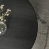 Czarno-drewniany, okrągły stolik kawowy z widoczną strukturą drewna, na którym stoi przezroczysty wazon z delikatnymi, jasnymi gałązkami. W tle widoczny jest fragment jasnego dywanu z ciemniejszymi wzorami.
