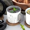 Porcelanowy kubek o pojemności 400 ml z szarym uchem, ozdobiony delikatnym wzorem kwiatowym, stoi na plecionym podkładce, wypełniony napojem z listkami mięty. Obok znajduje się drugi kubek z plasterkiem cytryny oraz fragmenty innych przedmiotów kuchennych.